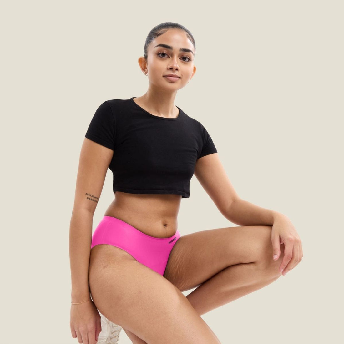 A confident young woman in a black crop top and bright pink WUKA period pants kneels against a neutral background. She has a calm, empowered expression and visible stretch marks, celebrating real bodies and period confidence.